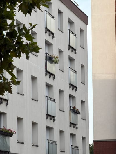 The image shows a close-up, side view of a modern apartment building in Maisons-Alfort, France, with its white facade punctuated by several balconies and windows. The building is tall, with multiple stories visible. The balconies are characterized by glass panels and dark metal railings, and some contain planters with flowers. Green foliage from a tree in the foreground partially obscures the upper left portion of the building, its leaves a dark green. Adjacent to the main building, on the right side of the frame, is another section of a building, a lighter beige color, with a dark red brick base visible at the very bottom. The sky is a pale, overcast blue. There are no people visible in the image, and the scene appears to be a still, quiet moment captured from an exterior perspective.