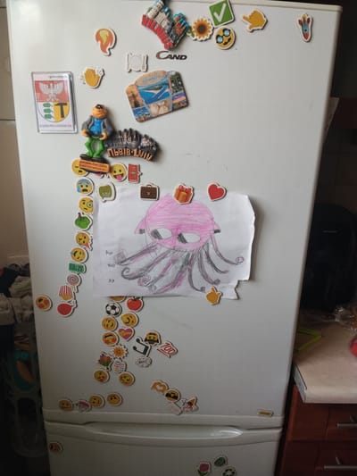 The image displays the front of a white refrigerator adorned with numerous magnets. Among the magnets are several emoji-style stickers, small souvenir magnets depicting cityscapes and landmarks, and a child's drawing of a pink octopus. One magnet is a coat of arms with the text "DĄBROWA GÓRNICZA" visible. The refrigerator is located in what appears to be a kitchen, with a countertop and cabinets visible to the right, and household items and laundry to the left. The scene is brightly lit, suggesting an indoor daytime setting. There are no people visible in the image.