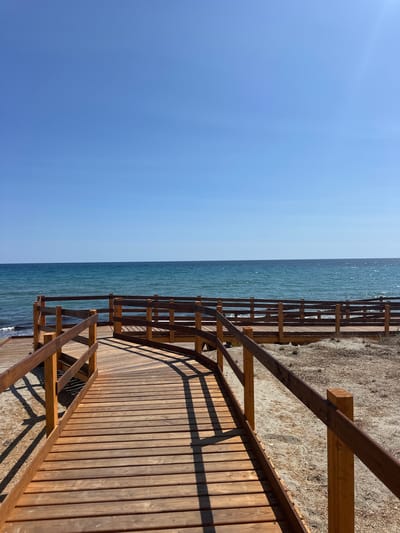The image shows a wooden boardwalk leading towards a calm blue sea under a clear blue sky. The boardwalk has wooden railings and casts shadows on the sandy ground. The sea appears tranquil with small waves gently lapping at the shore. The scene is set on a beach. There are no people or animals visible in the image. The location is the beach of Biddeputzi, Villaputzu, Italy.