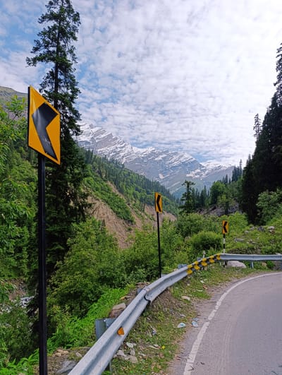 The image captures a scenic view of a winding mountain road in Dhundi, India. In the foreground, a metal guardrail with yellow and black hazard stripes lines the edge of the asphalt road. Several yellow directional signs, indicating a curve to the left, are visible along the roadside. Lush green trees and vegetation cover the steep slopes of the mountains. In the background, majestic snow-capped peaks rise against a sky filled with scattered white clouds. The overall impression is one of a remote and beautiful mountain landscape, typical of the region. There are no people visible in the image.