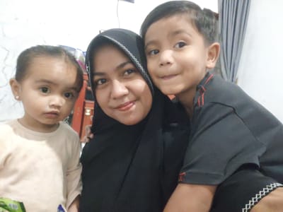 In this close-up selfie, a woman wearing a black hijab is positioned centrally, embracing a young boy who is on her right and a young girl who is on her left. The woman has a warm smile, with her tongue slightly visible. The boy, facing the camera, has dark hair and is wearing a dark t-shirt with red accents on the sleeves. The young girl, also looking towards the camera, has her hair pulled back and is wearing a light-colored long-sleeved shirt. She has a small pink earring in her left ear. The background is dimly lit, with a white textured wall on the left and grey curtains on the right. A hint of red furniture or a cabinet is visible behind the woman. The overall impression is a casual family moment captured indoors.