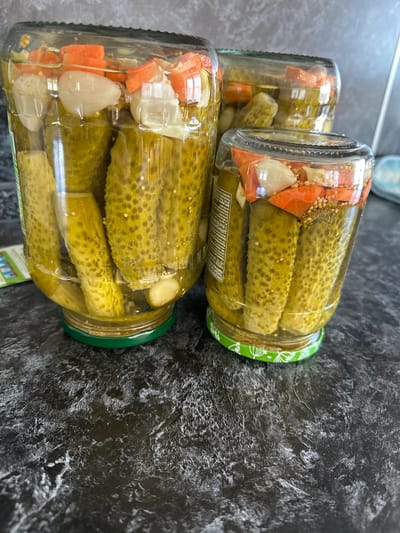 The image shows three glass jars of pickles on a dark, textured countertop. The jars are filled with whole pickles, brine, and visible pieces of chopped carrots and garlic. The jar in the foreground is the smallest and is slightly to the right of center. Behind it, to the left, is a taller, wider jar. A third jar is visible in the background, partially obscured by the other two. The jar lids appear to be metal, with the foreground jar having a green lid with a white floral pattern. The countertop has a marbled or stone-like appearance with lighter gray and white veining against a dark background. There are no people or animals present in the image, and no visible indication of the location being in Белозерки, Russia. The scene is static and focuses on the preserved food items.
