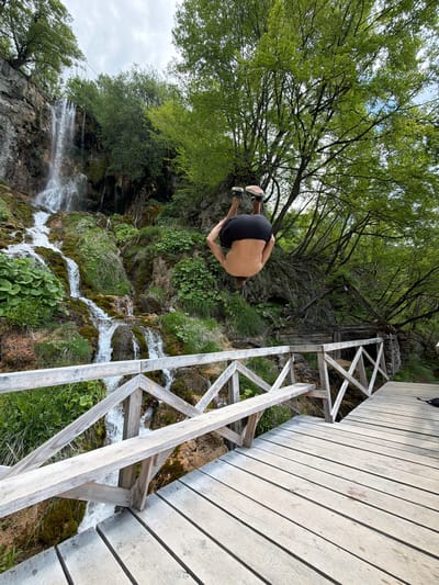 The image shows a man in mid-air, appearing to dive from a wooden platform near a waterfall. The man is wearing dark shorts and sneakers. He is positioned upside down with his head pointing towards the ground. The wooden platform is made of planks and has a wooden railing with a criss-cross design. Beneath the platform, water cascades down a mossy rock face, forming a waterfall. Green vegetation surrounds the waterfall. A tree with dense green foliage is situated above the man and waterfall. The scene is likely taken near the Sopotnica Waterfalls, which are located in the Prijepolje Municipality of Serbia.