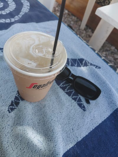 A close-up, high-angle shot captures a beverage in a Segafredo Zanetti branded cup with a clear plastic lid and a black straw. The cup is placed on a blue and white striped towel, which is situated on a pebbly surface, suggestive of a beach or outdoor seating area. Adjacent to the cup, a pair of black sunglasses rests on the towel. In the background, the legs of white outdoor chairs are visible, along with more of the pebbly ground. The lighting suggests an outdoor, daytime setting in the Municipal Unit of Diakopto, Greece, likely a cafe or beachside establishment, where one might enjoy a refreshing drink and relax.