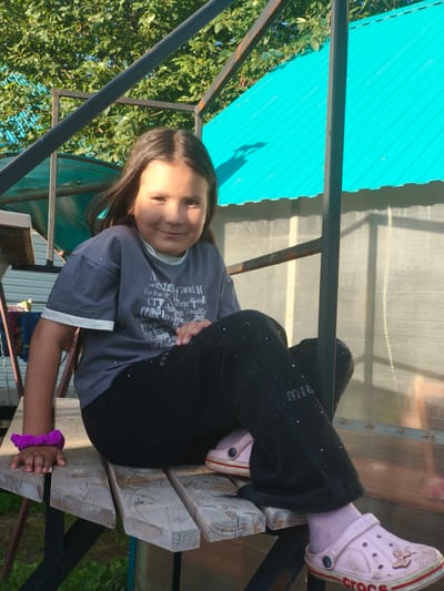 A young girl with dark hair and a light complexion is sitting on a wooden bench outdoors. She is wearing a dark grey t-shirt with a white trim, black pants with rhinestones, pink socks, and pink Crocs. The girl is smiling and looking at the camera. She has a purple scrunchie on her left wrist. The bench is part of a structure with a metal frame and a translucent corrugated plastic wall behind it. A bright teal-colored roof is visible above and to the right. Green foliage and trees are in the background. The scene appears to be in a residential yard, possibly in Burayevo, Russia, given the context. The lighting suggests it is daytime, with sunlight casting shadows.