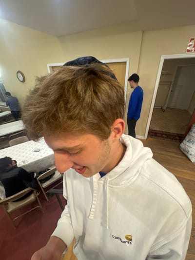 The image depicts an indoor scene in Bergenfield, United States, featuring four individuals. In the immediate foreground, a young male with light brown, wavy hair and visible facial stubble wears a white Carhartt hooded sweatshirt over a dark blue garment, with a dark cap positioned on the back of his head. He is positioned looking downward and slightly to his left, with his mouth partially open in what appears to be a smile or mid-speech.
In the mid-ground, to the left, another individual in a dark long-sleeved shirt is partially visible, leaning over a table covered with a white tablecloth. A dark object, possibly a smartphone, rests on this table. Folding chairs with light brown seats and dark frames are arranged around the table. A round, wood-framed clock with a white face is mounted on the light yellow wall behind this area.
Further back, to the right of the central figure, a third male in a royal blue long-sleeved shirt and dark pants stands facing away from the camera, partially framed by a white doorway. He wears a dark beanie. Beyond this doorway, another room with light wooden flooring and a dark patterned rug is visible, along with a partial red "EXIT" sign mounted high on the wall. A white bag with the text "DEPO" (likely "HOME DEPOT") is on the wooden floor near this doorway. The floor transitions from a red carpeted area on the far left to light wooden planks across the rest of the visible space. The walls are uniformly painted in a light yellow or cream color.