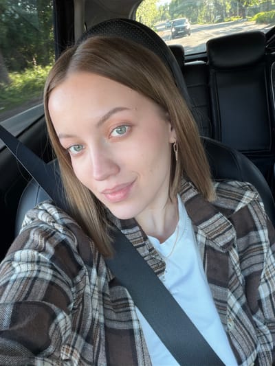 A woman with light brown hair and green eyes is in a car, wearing a white shirt and a brown plaid shirt. She is wearing a seatbelt. The location cannot be determined from the image provided. There is no indication of the city Maksimkovo, Russia.