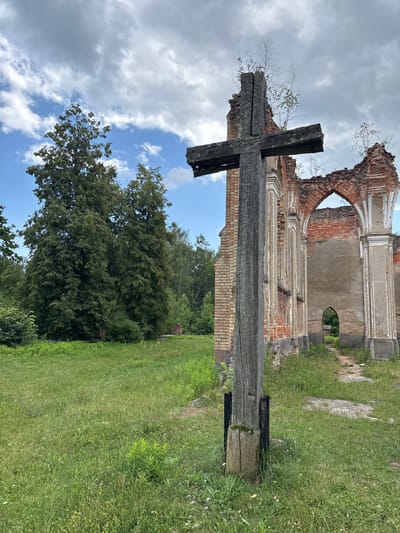 The image shows a wooden cross in the foreground, with the ruins of a brick building behind it. The building appears to be an old church, with arched windows and weathered brickwork. Trees and greenery surround the ruins, suggesting an outdoor, possibly rural, location. The sky is cloudy. The scene is set in the city of Bachury, Poland. There are no people visible in the image.