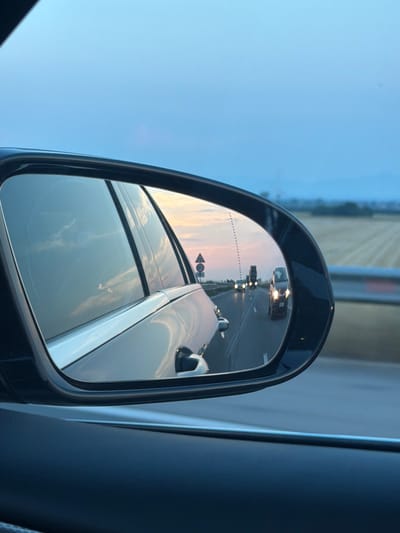 An image captured in Mirovo, Bulgaria, depicts a view from the interior of a vehicle, primarily focusing on its driver's side rear-view mirror and the external environment at what appears to be dusk. The foreground of the image features the dark trim and interior paneling of the car, with the large, dark-framed side-view mirror prominently positioned on the right.

Reflected within the mirror is a multi-lane highway, with traffic moving in the same direction as the observer. Several vehicles are visible, all with their headlights illuminated. The closest reflected vehicle, a compact car or utility vehicle, faces the observer directly, its headlights distinct. Further down the highway, additional vehicle lights are discernible. A dashed line, indicative of a blind spot monitoring system or a mirror warning area, is present on the left side of the mirror's reflection. A triangular road sign with a red border and a black symbol is also visible on the roadside within the reflection. The reflected sky above the highway displays a gradient from light blue to soft pink and orange hues, characteristic of twilight.

Beyond the mirror's frame, on the right side of the primary image, a wide, flat agricultural field with visible crop rows extends towards the horizon. A barrier or guardrail runs along the right edge of the highway. The overarching sky, visible above the car's structure and the distant landscape, is a clear, pale blue, transitioning subtly towards the horizon. The environment suggests a rural or semi-rural setting alongside a well-maintained highway. No people are explicitly visible in the scene.