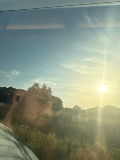 This is a selfie taken outdoors, likely during the day, as indicated by the bright sunlight and lens flare. The subject is a man with a beard and possibly short hair, his face partially visible on the right side of the frame. He appears to be looking to his left, with his eyes closed or downcast.
The background is a bright, hazy sky with scattered clouds, suggesting fair weather. The sun is prominently featured, creating a strong glare that washes out some details. On the right side of the image, there is a suggestion of foliage or trees, with a dappled, green-brown texture.
The image is oriented vertically, with the subject's head towards the top and the sky filling much of the frame. There is a reflection on what appears to be a glass surface on the left side of the image, showing a light gray, possibly vertical structure, and a muted reflection of the sky. This reflection, along with the overall lighting, suggests the photo might have been taken through a window or windshield.
No text is visible in the image. The overall mood is somewhat relaxed and perhaps contemplative, with the soft focus and natural lighting contributing to this impression. The location context of Wuustwezel, Belgium, does not provide specific visual cues within the image itself.