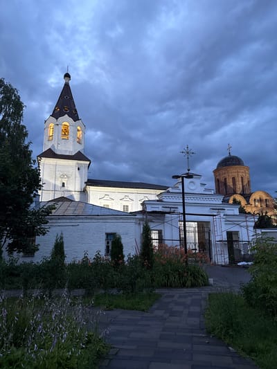 This image captures a view of a church, likely the Church of the Resurrection of Christ in Demenshchina, Russia. The perspective is tilted, with the church structure dominating the frame from the bottom left up to the top right.
The church features white brick walls with dark roofing. A prominent bell tower, also white with a dark roof and a spire, is visible on the left. The tower has arched windows that are lit from within, suggesting an evening or nighttime setting. Further into the image, a larger dome structure of the church is visible, made of reddish-brown brick with a dark dome. A cross stands atop the dome. The church appears to be partially undergoing renovation, with scaffolding visible on the right side of the main building.
The foreground and right side of the image are filled with lush greenery and landscaping. A paved walkway made of grey bricks runs along the right edge of the church, bordered by dense bushes and flowering plants, some with small orange blooms, and others with delicate purple flowers.
The sky is a dramatic, cloudy grey, indicating either dusk, dawn, or overcast weather. The lighting on the church, particularly the illuminated windows, suggests it is either twilight or nighttime. No people are visible in the image. There is no visible text in the image. The overall mood is serene and a little somber due to the overcast sky and the hints of renovation.