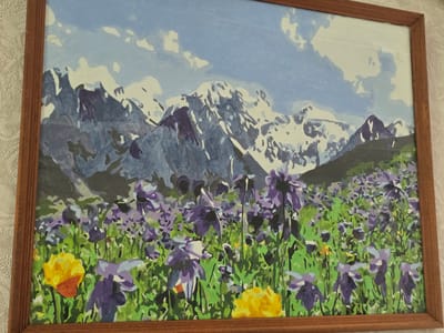 A framed painting depicts a mountain landscape with a field of flowers in the foreground. The sky is a light blue with scattered white clouds. Snow-capped mountains with dark purple and gray peaks dominate the background. The foreground is filled with lush green grass and numerous purple flowers, with two bright yellow flowers and one red and yellow flower in the lower portion of the field. The painting is enclosed in a simple wooden frame. The location is not discernible from the image.