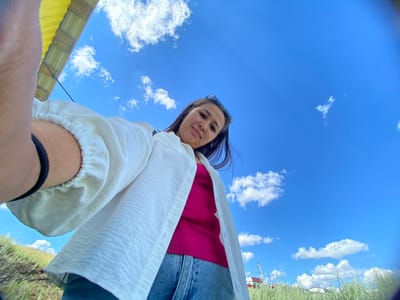 A low-angle, wide-angle shot captures a young woman looking up at the camera from below, framed against a bright blue sky with scattered white clouds. She is outdoors, possibly in Yuldybayevo, Russia, given the grassy hillside and distant structures. The woman has long, dark hair and is wearing a white textured shirt over a pink ribbed tank top and blue jeans. Her left arm is raised, holding the camera, with a black elastic band visible on her bicep. In the background, a yellow structure with visible wooden beams is partially visible at the top left of the frame. The landscape features dry, green grass and a few small buildings in the distance, with power lines visible to the right. The overall impression is one of a casual, outdoor moment.