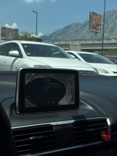 The image captures a detailed view from inside a vehicle, looking out onto a road scene in Ciudad Santa Catarina, Mexico.
In the foreground, a dark, textured vehicle dashboard is prominent. Central to the dashboard is a digital infotainment screen, which exhibits significant concentric cracking, particularly around its perimeter, radiating from a central dark area. The visible text on the screen includes "R-FM," "FT JORGE MEDIN," and "INA DEL TIEMPO P 40." Below the screen are horizontal air vents and a red, triangular hazard light button. A small, round, red air freshener labeled "ewlys" is suspended from the rightmost air vent.
The midground reveals several white passenger vehicles ahead, suggesting traffic or a stop on a multi-lane road. In the background, large, light-colored mountains dominate the horizon, characteristic of the geography surrounding Ciudad Santa Catarina. An elevated concrete overpass spans the scene horizontally between the vehicles and the mountains. Two billboards are visible, one partially obscured on the left, and another more distinct on the right displaying an advertisement. The environment is illuminated by bright sunlight under a clear blue sky with scattered white clouds.