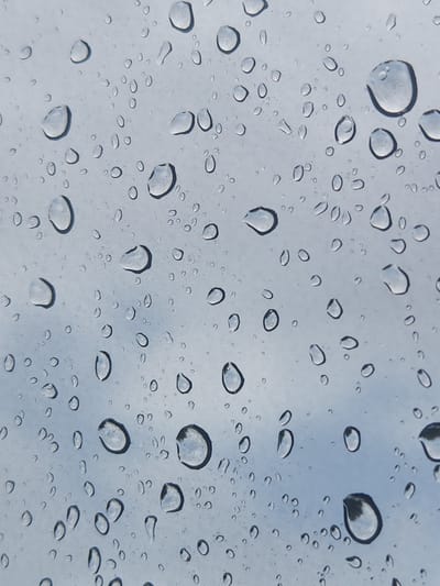 A close-up, high-angle photograph shows a surface covered in numerous water droplets of varying sizes. The droplets are scattered across a light blue, overcast sky or a similar reflective surface. The light refracts through the droplets, creating highlights and dark outlines, giving them a three-dimensional appearance. Some droplets are perfectly spherical, while others are elongated or irregularly shaped, suggesting movement or surface tension. The overall impression is of a rainy day, with water accumulated on a window or another smooth, transparent surface. The image is devoid of any human presence or urban elements, focusing solely on the natural phenomenon of water droplets.