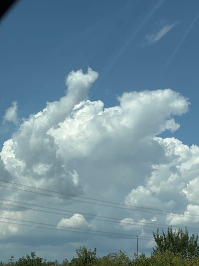 The image depicts a cloudy sky with power lines and trees in the foreground. A large cumulus cloud formation dominates the center of the frame, with smaller clouds scattered across the blue sky. Sunlight beams are visible, suggesting it is daytime. The power lines are strung across the lower portion of the image, and a dense line of trees and foliage forms the base of the scene. The context of Chiasottis / Cjasotis, Italy is not present in the visual information of the image.