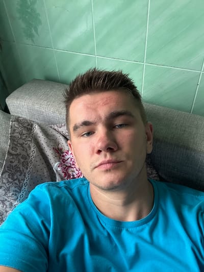 A young Caucasian male is shown from the chest up, resting against a grey couch with a patterned textile. The male has short, spiked brown hair, blue eyes, and is wearing a bright blue t-shirt. He has light stubble on his face and appears to be looking directly at the camera. Behind him, a wall is visible, tiled in a light green color. The lighting suggests an indoor environment, possibly a home. No specific location in the city of Krasnoe, Russia, can be identified from the image.