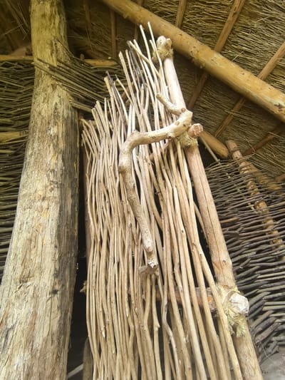 The image shows the interior of a rustic structure, possibly a hut or shelter, constructed primarily from natural materials such as wood and branches. A large, rough-hewn wooden post stands prominently on the left. To its right, a vertical barrier or doorway is formed by a dense arrangement of slender, light-colored branches, some of which appear to be tied together. A gnarled, bent branch extends horizontally across the upper portion of this branch barrier, adding to the organic and handmade aesthetic. In the background, to the right, a woven wall or screen made of darker, thinner branches is visible. The ceiling is supported by wooden beams and covered with a thatched material, likely straw or reeds, creating a textured, rustic roof. The overall impression is one of simple, traditional construction, suggesting a rural or historical setting. The location of Камянюцкі сельскі Савет in Belarus is noted as the context for this structure. There are no people or animals visible in the image.