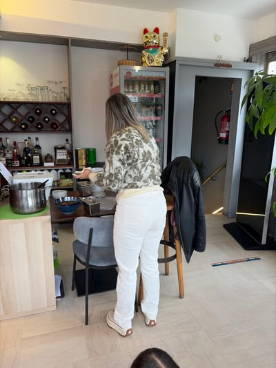 This image presents an interior scene within a commercial establishment, likely a restaurant or cafe, located in Walferdange, Luxembourg.
A female individual stands in the central foreground, her back to the camera. She wears a cream-colored sweater with a brown and green abstract pattern, white trousers, and white platform shoes. Her hands are positioned over a counter, appearing to be handling a small white object.
To the left, a bar area features upper shelves displaying inverted wine glasses and a lower wooden wine rack containing multiple bottles. Below this, various liquor bottles are arranged on a counter surface, alongside a small, upright black container with Asian script. A silver pot rests on a green placemat on a light wooden cabinet.
In the mid-ground, behind the standing individual, a refrigerated display cooler, branded with 'Coca-Cola', is visible, stocked with assorted beverages. A golden Maneki-neko (waving cat) figurine and a miniature Japanese umbrella are positioned on top of this cooler. Below the cooler, a wooden table holds a blue bowl containing a dark substance and several square metal containers. A gray chair and a wooden stool with a black leather jacket draped over it are also present.
To the right, a doorway leads to a darker area, revealing a red fire extinguisher mounted on the wall and a handrail indicating a staircase. A large green plant is partially visible on the far right. The floor is covered with light-colored, square tiles, and a multi-colored linear graphic, resembling a ruler, is visible on the floor in the mid-right. A portion of a dark-haired head is visible in the lower foreground, indicating the presence of another individual at a lower vantage point.