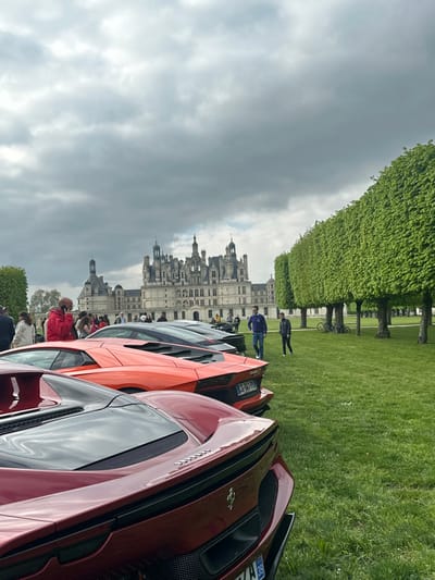 The image shows a red Ferrari parked in front of a bright orange Lamborghini. The Lamborghini is parked in front of a black car. The cars are parked in a grassy field. The Chateau de Chambord, a large castle in the Loire Valley, is visible in the background. The castle is made of white stone and has many towers and turrets. It is surrounded by a moat. The sky is cloudy. There are a few people walking around. The coordinates provided, 47.615 latitude and 1.519 longitude, place the scene in the Chateau de Chambord's grounds in the city of Chambord, France.
