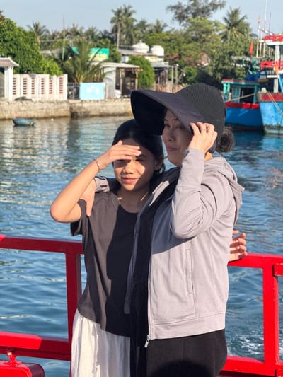 A woman and a girl are standing on a red railing, possibly on a boat or dock, in the city of An Hai, Vietnam. The woman is wearing a gray hooded jacket and a black hat, while the girl is wearing a dark t-shirt and a white skirt. Both are looking towards the water. In the background, there are buildings, palm trees, and a blue boat. The water is choppy and reflects the sunlight. The scene appears to be during the daytime.