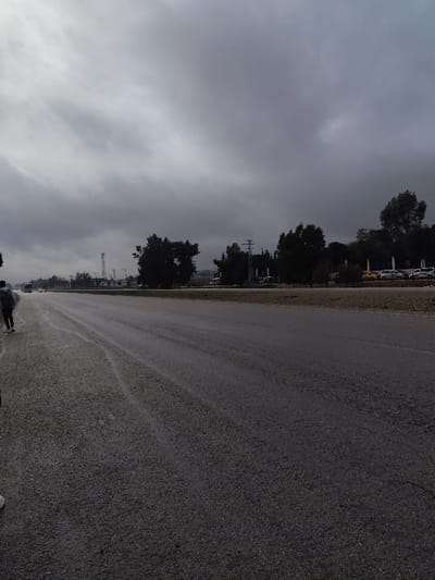 The image depicts an outdoor scene in Mankat al-Hatab, Syria, under a uniformly overcast, dark grey sky. A wide, multi-lane asphalt road, visibly wet and textured, extends from the foreground into the distance, occupying the majority of the lower and central frame. On the far left, a person wearing dark clothing and a backpack is observed walking away from the viewer on the road's shoulder. A white shoe, belonging to a second individual, is partially visible at the extreme bottom left corner. To the right of the asphalt road is a narrow, dry grass median strip. Beyond this median, a dense line of trees with dark foliage runs parallel to the road. In the background, behind the trees, a prominent utility pole with multiple cross-arms and a slender lattice telecommunications tower are visible. Further right and behind the tree line, an area containing multiple parked vehicles, including a distinct yellow car, can be identified. The distant horizon reveals faint outlines of hills or structures, largely obscured by the heavy cloud cover and subdued lighting.