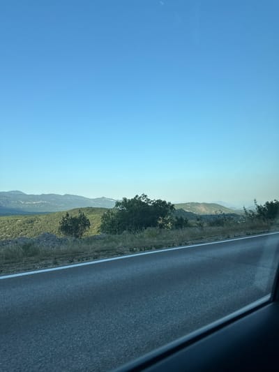 The image depicts a landscape scene from the perspective of a vehicle traveling along a road. The road itself, paved with asphalt, occupies the right side of the frame and features a white lane marker. To the left of the road, a steep, green, and rocky hillside rises, dotted with trees. The hillside recedes into the distance, forming rolling mountains under a clear, bright blue sky. The sun is high, suggesting it is daytime. The scene is natural and undeveloped, with no signs of human structures other than the road. The image conveys a sense of travel and scenic beauty, possibly experienced from a road trip through a mountainous region.