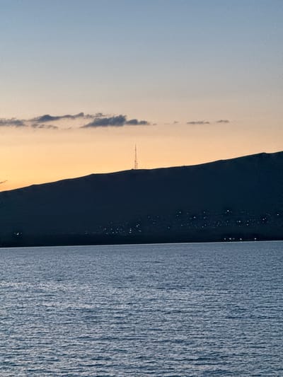 The image depicts a panoramic view of Tsamakaberd, Armenia, during twilight. The foreground is dominated by a body of water, likely a lake or reservoir, with rippling surface reflecting the fading light. In the mid-ground, a dark, silhouetted mountain range forms the backdrop. Along the base of the mountain, a scattering of lights indicates a settlement or town. A prominent tower, likely a telecommunication mast, stands on the crest of the mountain, piercing the sky. The sky above is a gradient of colors, transitioning from a pale orange near the horizon to a soft blue at the top, with a few wispy clouds present. The overall scene is serene and captures the tranquil ambiance of the location at dusk.