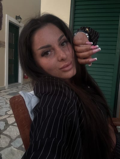 The image is a close-up, eye-level shot of a woman with dark hair and freckles, likely taken in the Municipal Unit of Lefkada, Greece. She is wearing a black pinstripe blazer and has long, manicured nails with a white tip design. Her hand is resting on her face, and she appears to be looking directly at the camera. Behind her, there is a green shuttered window, a beige exterior wall, and a glimpse of stone tile flooring. A wall mounted exterior light can be seen attached to the beige wall.