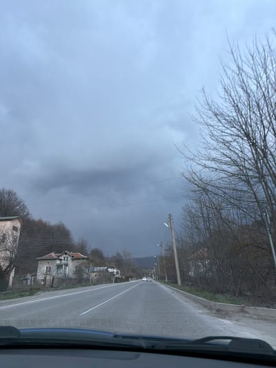 The image depicts a road in the village of Gintsi, Bulgaria, under a heavily overcast sky. The road is wet, likely from recent rain. A white dividing line runs down the center of the road. On the right side of the road are houses, most appearing somewhat dilapidated. Trees, bare of leaves, line the roadside. Utility poles and wires are visible along the road. The view is likely taken from inside a vehicle, as part of a dashboard is visible on the right side of the frame. No people are visible. A couple of cars appear far off down the road, but they lack detail.