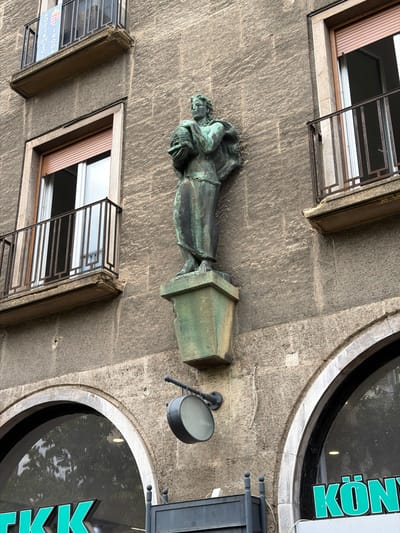 This is a low-angle, medium shot of a bronze sculpture of a human figure on the exterior of a building in Debrecen, Hungary. The sculpture is green with patina and depicts a person holding a round object, possibly a beehive, in their arms. The figure stands on a weathered green pedestal that is mounted on the building's stone facade.
Above the sculpture, there are two windows with balconies. The left window has a rolled-down brown shutter, and the right window shows an open shutter revealing a white interior. Both balconies have black metal railings and stone ledges, with the left ledge showing signs of wear and damage. A banner with Hungarian colors and text hangs from the left balcony.
Below the sculpture and to the left, part of a large, arched window or storefront is visible, with the green neon letters "TKK" illuminated. To the right of the arched window, the green neon letters "KÖNY" are visible on another storefront. A circular, dark-colored sign hangs from an articulated arm on the building's facade between the sculpture and the arched windows. The building's facade is constructed of rough-hewn stone with visible horizontal striations. The overall impression is of an urban architectural setting.