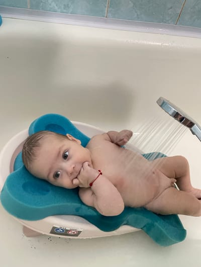 The media file depicts a baby being bathed in a bathtub. The baby is lying on a blue bath support, which is placed in a white bathtub. A shower head is visible at the top left of the image, with water spraying down onto the baby. The baby has light brown hair and is looking at the camera with a curious expression. The baby is wearing a red bracelet on its left wrist. The background shows a portion of a tiled wall in a light blue color. There are some water droplets on the surfaces. A sticker is visible on the side of the bath support, with some text and symbols. One symbol appears to be a warning sign. Another symbol looks like a checkmark within a circle. The word "MAX" is also visible on the sticker. The scene appears to be indoors, in a bathroom. The lighting suggests it could be daytime.