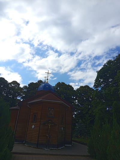 The image captures a wooden Orthodox church with a blue dome and a golden cross, set against a backdrop of lush green trees and a cloudy sky. The church, located in the city of Giaginskaya, Russia, has a rustic wooden facade with visible horizontal planks. A crucifixion scene is mounted on the exterior wall, featuring a golden crucifix with Jesus and inscriptions. The church has a red-tiled roof and several windows. The surrounding environment is characterized by dense foliage, including evergreen trees and bushes, and a paved walkway leading towards the church. The sky is a mix of blue and white clouds, suggesting a partly cloudy day. No people are visible in the image.