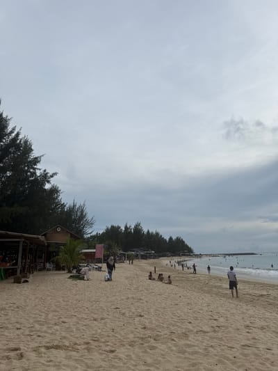 The image depicts a sandy beach with several people present. The sky is overcast with scattered clouds. On the left side of the image, there are some structures that appear to be wooden huts or stalls, with people visible inside some of them. Trees line the background behind these structures. Further down the beach, there are more people scattered along the sand and in the water. Some individuals are walking, while others are sitting or playing. The ocean is visible in the distance, with a breakwater or pier extending into the water on the far right. The sand is a light brown color, and there are footprints visible in some areas. The scene suggests a public beach with some amenities.