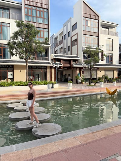 A young woman with dark hair, wearing a brown strapless top and a white skirt, stands on a large circular stepping stone in a shallow body of water. She is wearing glasses and white sneakers. The water reflects the surrounding buildings. In the background, there are modern, multi-story buildings with large windows and balconies, some adorned with wooden slats. Trees and potted plants are visible along the walkways and in front of the buildings. A yellow origami crane sculpture floats in the water to the right. Signs on the buildings indicate commercial establishments, with one reading "KOREA BINSU". The scene appears to be an outdoor public space within a planned urban development in Nghĩa Trụ Commune, Vietnam, possibly a commercial or residential complex. The lighting suggests it is daytime, with clear skies and some shadows.