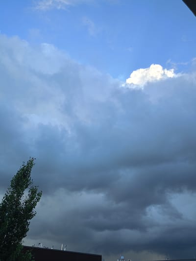 The image depicts a scene of dramatic skies over Peetri alevik, Estonia. The sky is filled with dark, heavy storm clouds dominating the frame, with a hint of brighter blue and a single, bright white cloud patch visible in the upper right quadrant. A tall, leafy green tree stands in the lower left, its branches reaching towards the turbulent sky. Below the tree and clouds, the dark roofline of a building is visible, featuring several metal chimney-like structures. The overall impression is of an impending storm.