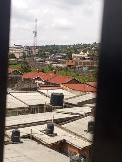 The image displays an elevated view of a densely developed urban landscape in Kiamaina ward, Kenya, observed through a vertical opening, indicated by dark vertical bars on the left and right edges.

In the immediate foreground, multiple rooftops constructed from corrugated silver-grey metal are visible. These roofs are relatively flat or gently sloped. Several black cylindrical plastic water tanks, labeled "POA TANK," are mounted on wooden or concrete supports on these roofs. Partial views of the concrete and stone walls of the buildings below are also discernible.

The midground features numerous residential structures with predominantly red or orange corrugated metal or tiled roofs. These buildings are interspersed with stone walls, overgrown grassy areas, and small trees. Beyond these houses, the terrain rises, revealing more varied architecture, including multi-story buildings of concrete and brick construction.

The background is dominated by a tall, lattice-style telecommunications tower, painted white and red, situated on slightly elevated ground. Further in the distance, a cluster of multi-story buildings, some appearing commercial or residential, is visible. The furthest background shows a hilly landscape covered with dense green vegetation, indicating natural or undeveloped areas. The sky is overcast with white and grey clouds. No individuals or specific actions are discernible within the frame.
