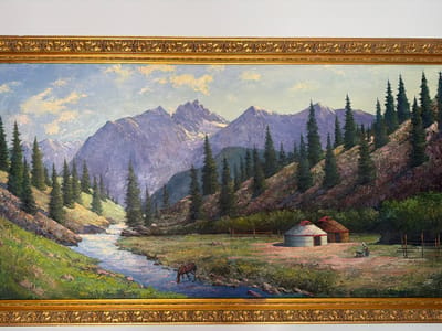 This is a wide-format oil painting on canvas, framed in an ornate gold-leaf gilded wood frame, depicting a serene mountain landscape. The scene features snow-capped purple and gray mountains in the background, under a light blue sky with scattered white and yellow clouds. In the middle ground, a winding, clear blue river flows from left to right, bordered by lush green grassy hills and numerous dark green pine trees. On the right side of the river, in the foreground, are two traditional nomadic dwellings, possibly yurts, one white with a red-topped roof and the other brown. A wooden fence encloses a small area. A solitary brown horse is drinking from the river on the left side of the painting. A woman in traditional clothing is depicted near the dwellings, seemingly engaged in some activity. The overall impression is one of a peaceful, remote, natural environment. The painting is displayed in an indoor setting, against a plain white wall, consistent with an art exhibition or gallery. The location is identified as Ankara, Turkey.