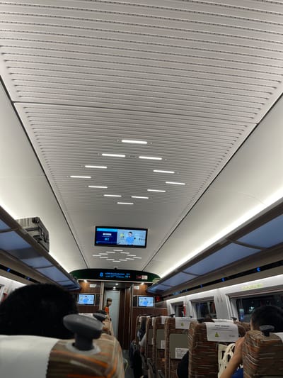 This image depicts the interior of a train carriage. The ceiling is made of white, perforated panels with linear light fixtures. A screen mounted on the ceiling displays information, including "2026-06-26", "16:38", and Chinese text that appears to indicate a destination or announcement. There are seats with striped upholstery visible, and some passengers are seated. Luggage is stored overhead. The overall environment suggests a modern, clean, and functional train interior. The city of Longhua District, China, is mentioned, but there is no direct visual evidence in the image to confirm this specific location.