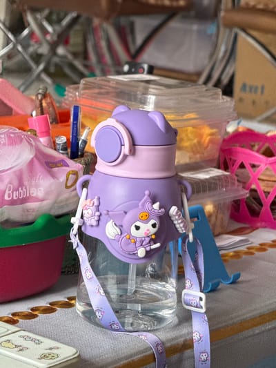 The image shows a purple water bottle with a character on it, possibly a cartoon character. The bottle has a strap with the same character on it. The bottle is on a table with other items, including a pink basket and some plastic containers. There is also a pink pack of tissues or wipes on the table, labeled "Soft Bubbles." In the background, there are shelves with various items, and some of the items are out of focus. The lighting suggests it is daytime. The image does not provide any specific location information within Manzhouli, China.
