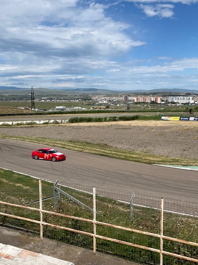 A red racing car travels along the Rustavi International Motorpark track in Georgia. The background displays the city of Rustavi under a partially cloudy sky. A metal and wire fence runs parallel to the track's edge.
