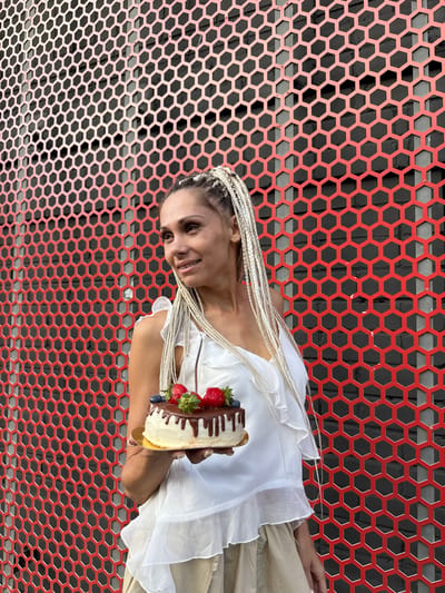 A woman with blonde braided hair stands in front of a red, honeycomb-patterned metal fence. She is holding a small cake topped with chocolate icing, strawberries, and blueberries. She is wearing a white ruffled top and a light-colored skirt. The background appears to be an outdoor urban setting.