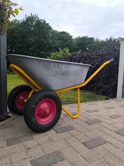 A grey wheelbarrow with a yellow frame and pink wheel hubs sits on a paved walkway. Behind the wheelbarrow is a dark, leafy bush, and beyond that, green trees. The sky is overcast. The wheelbarrow is positioned facing left, with its handles extending towards the right side of the frame. The paving stones are rectangular and laid in a herringbone pattern. A dark grey post is visible to the left of the wheelbarrow.