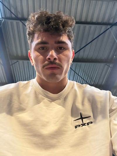 A male individual is depicted in a close-up, chest-up shot within an indoor environment in Chantepie, France. The subject, with dark, curly hair and a mustache, faces directly towards the camera. He wears a light-colored, long-sleeved sweatshirt featuring a black embroidered logo on the upper left chest, consisting of a cross-like symbol above the letters "PXP." A small silver hoop earring is visible in his left earlobe. The background consists of a corrugated metal ceiling structure, consistent with an industrial or commercial building interior, with dark support beams visible.