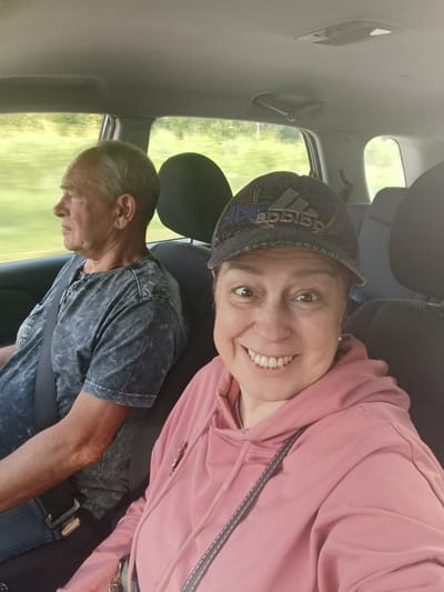 Two people are in the front seats of a car, traveling down a road. The person on the right is a woman taking a selfie, smiling and wearing a pink hooded sweatshirt and a dark baseball cap with the Adidas logo. She has brown eyes and is looking at the camera. The person on the left is a man wearing a gray, distressed-looking t-shirt and a seatbelt. He is looking out the car window to his left, with a neutral expression. The background outside the car window shows a blurred green landscape, suggesting motion and a daytime setting. The interior of the car is visible, including the car's ceiling, headrests, and seatbelts. There is no visible text in the image. The scene appears to be a casual road trip in a rural or suburban area, given the greenery outside. The lighting suggests it is daytime, possibly overcast or late afternoon given the soft light.