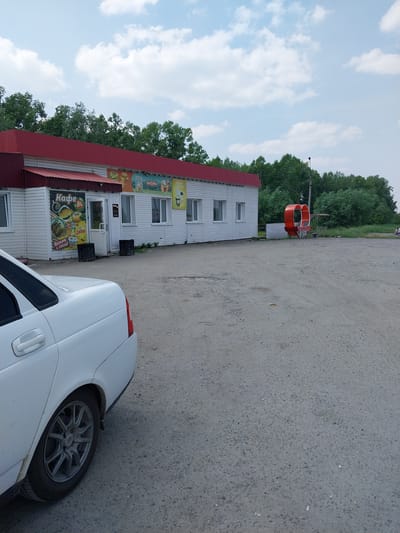 This is an outdoor, eye-level, slightly tilted, landscape photograph taken on a bright, cloudy day in Troitskoe, Russia. The scene features a small, white, single-story building with a red roof, likely a cafe or small shop, with several windows and signs on its exterior. One prominent sign in Russian reads "Кафе 24" which translates to "Cafe 24". Other signs depict food items and drinks, including coffee. In front of the building, a white car is parked, with its front wheel visible. Behind the building and to the right, a patch of pavement is visible, as are trees in the background. To the left of the building, there is a distinctive bright orange, circular structure, possibly a part of a playground or amusement ride. The sky is blue with scattered white clouds. The overall impression is a simple, functional roadside establishment on a typical day.