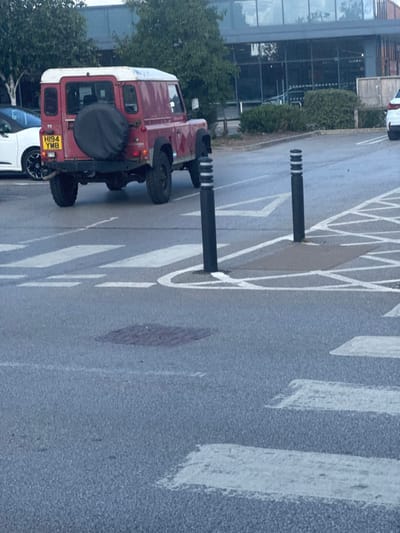 This image depicts a car park scene in Pocklington, United Kingdom, adjacent to a large, modern retail building, consistent with the Sainsbury's supermarket on Broad Street.
A red Land Rover Defender, with a white roof and a black spare wheel cover mounted on its tailgate, is the primary vehicle subject, viewed from the rear in the mid-ground. Its UK registration plate, "HI94 YMB," is clearly visible. The vehicle appears to be angled as if in the process of turning or recently stopped. To the immediate left, a portion of a white modern vehicle is partially visible, parked. The car park surface is asphalt, appearing slightly damp, with clearly defined white painted markings including sections of a zebra crossing in the foreground and diagonal hatched lines further back. Two black bollards, each with two reflective white bands, stand vertically in the mid-foreground. Two circular manhole covers are embedded in the asphalt: one in the foreground, and another further back to the right. In the background, the large, multi-story commercial building features extensive dark-tinted glass windows that reflect the overcast sky. Shopping trolleys are faintly visible through these windows. Green foliage, including trees and bushes, is present around the building and car park perimeter. No individuals are visible within the frame.
