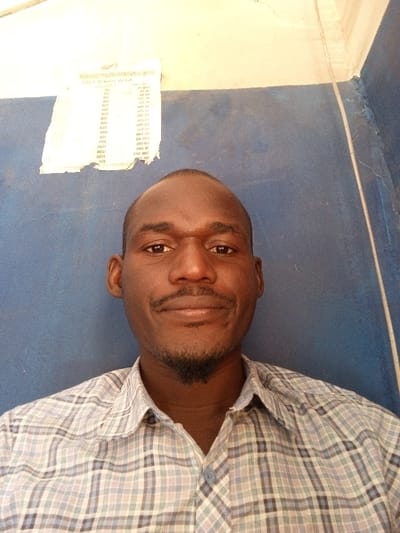 A male individual is depicted in a head-and-shoulders, frontal portrait within an interior setting in Chiranchi, Nigeria. He possesses dark skin, a bald head, a mustache, and a short, pointed goatee. He wears a light-colored plaid button-down shirt with blue, grey, and white patterns and is looking directly at the camera with a neutral expression. The background features a two-toned wall: an upper, light beige/off-white section meeting a lower, dark blue section. In the upper-left quadrant, a crumpled, rectangular piece of paper is affixed to the wall, partially overlapping the beige and blue sections. Text on the paper, including "NEW PRICE LIST" and numerical entries, is visible and appears inverted, suggesting it might be a reflection or a reversed image of the original text. Cracks are visible where wall sections meet in the upper right. No other individuals or objects are present, and no specific actions or interactions are discernible beyond posing for the photograph.