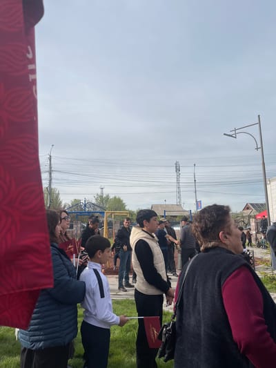 A photograph taken in Akhuryan, Armenia shows a group of people, predominantly children and adults, lined up outside, potentially for an event or ceremony. Many are holding small flags, the majority of which have a red background and an emblem. The environment includes utility poles, power lines, and a telecommunications tower. In the background, a fence can be seen. The sky overhead is overcast. The individuals are wearing a variety of clothing, ranging from jackets to sweaters. Some are looking in the same direction, possibly towards a focal point of attention. A section of a large red banner with lettering is visible in the foreground.