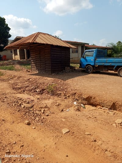 The image shows a dirt road in front of a small wooden hut with a corrugated metal roof. The hut is located in Ilobu, Nigeria, at latitude 7.836 and longitude 4.476. A blue pickup truck is parked to the right of the hut. The road is rough and dusty and there is a small ditch beside it. The environment is dry and arid. The sun is shining brightly and there are no people in the image. The scene appears to be in a rural area.